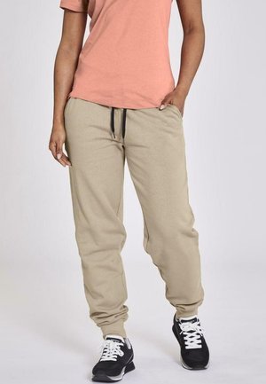 Beige joggings met een trekkoord in de taille, elastische boorden en een zachte textuur, gecombineerd met een perzikkleurig T-shirt en zwarte sport schoenen.