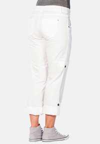 Białe spodnie cargo z podwiniętymi nogawkami, tylnymi kieszeniami i akcentami guzika. Noszone z szarymi sneakersami za kostkę. Gładka tekstura materiału.