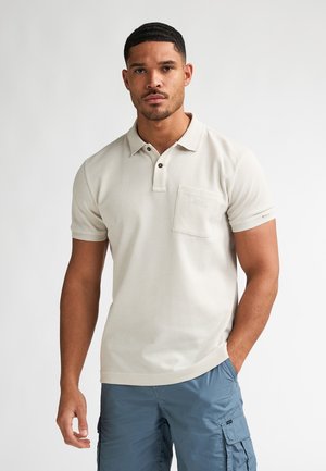 Roomkleurig polohemd met korte mouwen, een gestructureerde stof, een knoopsluiting met drie knopen en een borstzak. Draagt lichtblauwe cargoshorts.