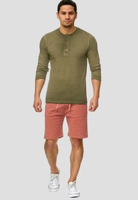 Chemise henley à manches longues verte olive avec trois boutons, associée à un short rose et des baskets blanches, illustrant un look décontracté d'été.