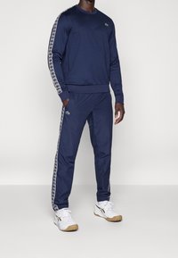 Námořnický modrý teplákový komplet vyrobený z měkké látky, s klasickým kulatým výstřihem, elastickými manžetami a vzorovanými postranními pruhy. V kombinaci s bílými teniskami.