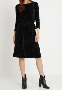 Vestido negro de manga larga con un diseño texturizado y plisado, y un cinturón en la cintura. Combinado con botines negros de tacón con detalle de cremallera lateral.
