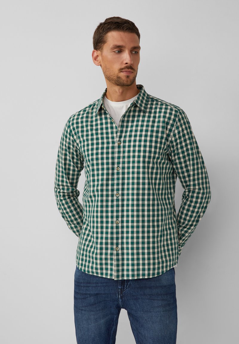 Man in een groen en wit geblokt overhemd met knopen over een wit T-shirt, staand met de handen achter zijn rug, kijkend naar links tegen een neutrale achtergrond.