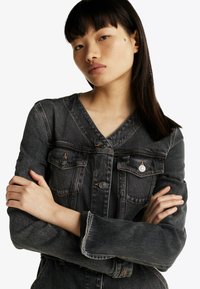 Giubbotto in denim nero con scollo a V, maniche lunghe, tasche sul petto e bottoni argentati. Presenta una texture leggermente consumata e una vestibilità casual.