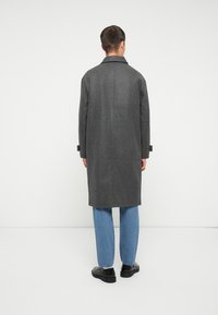 Grijze wollen overjas met een geknipte kraag en lange mouwen. Bevat zijsplitten en minimale details, gecombineerd met lichtblauwe jeans en zwarte schoenen.