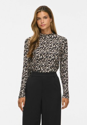 Jeune femme aux longs cheveux ondulés portant un haut à manches longues noir et beige à motifs, rentré dans un pantalon noir taille haute, debout sur fond blanc.