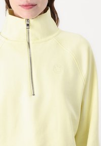 Helles, gelbes Sweatshirt mit hohem Kragen und silbernem Reißverschluss, das ein kleines Logo auf der Brust und einen weichen, strukturierten Stoff aufweist.