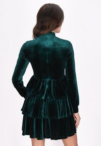 Rochie din catifea verde smarald, cu mânecă lungă, design cu volane în straturi și corset ajustat, evidențiind o textură moale și o finisare netedă.