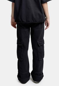 Personne debout portant un pantalon cargo noir avec de grandes poches latérales et un haut noir, vue de dos sur un fond uni.