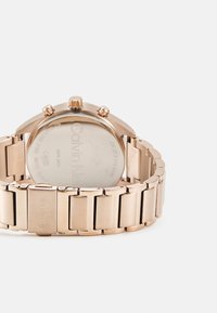 Montre en acier inoxydable rose gold avec un bracelet métallique, boîtier rond, trois poussoirs et un dos en verre lisse montrant le nom de la marque.