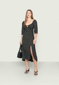 Vestido preto com padrão floral, mangas bufantes até o cotovelo, decote aberto e uma fenda lateral. Modelo combinou com uma bolsa preta e sapatos de salto.