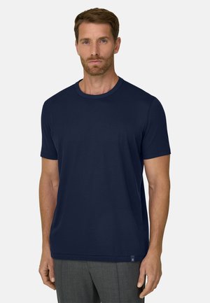 Homme aux cheveux courts bruns et barbe, portant un t-shirt bleu marine uni à manches courtes et un pantalon gris, debout sur un fond blanc.