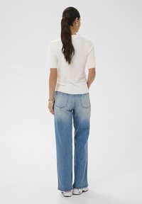 Femme aux longs cheveux bruns attachés en queue de cheval, portant un haut blanc à manches courtes, un jean large bleu et des baskets blanches, debout de dos.