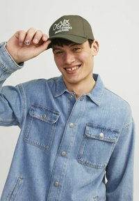 Gorra verde oliva con logotipo bordado en blanco, llevada por una persona con una chaqueta de mezclilla clara que tiene dos bolsillos en el pecho y botones de presión plateados.