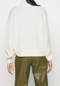 Maglione bianco lavorato a maglia con collo alto, polsini e bordo a costine; presenta una vestibilità ampia e cuciture delle spalle abbassate. Abbinato a pantaloni verde oliva.
