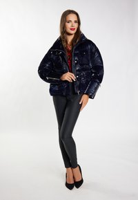 Marineblaue Steppjacke mit glänzender Oberfläche, ausgestattet mit mehreren Taschen und einem hohen Kragen. Kombiniert mit schwarzen, taillierten Hosen und schwarzen Pumps.
