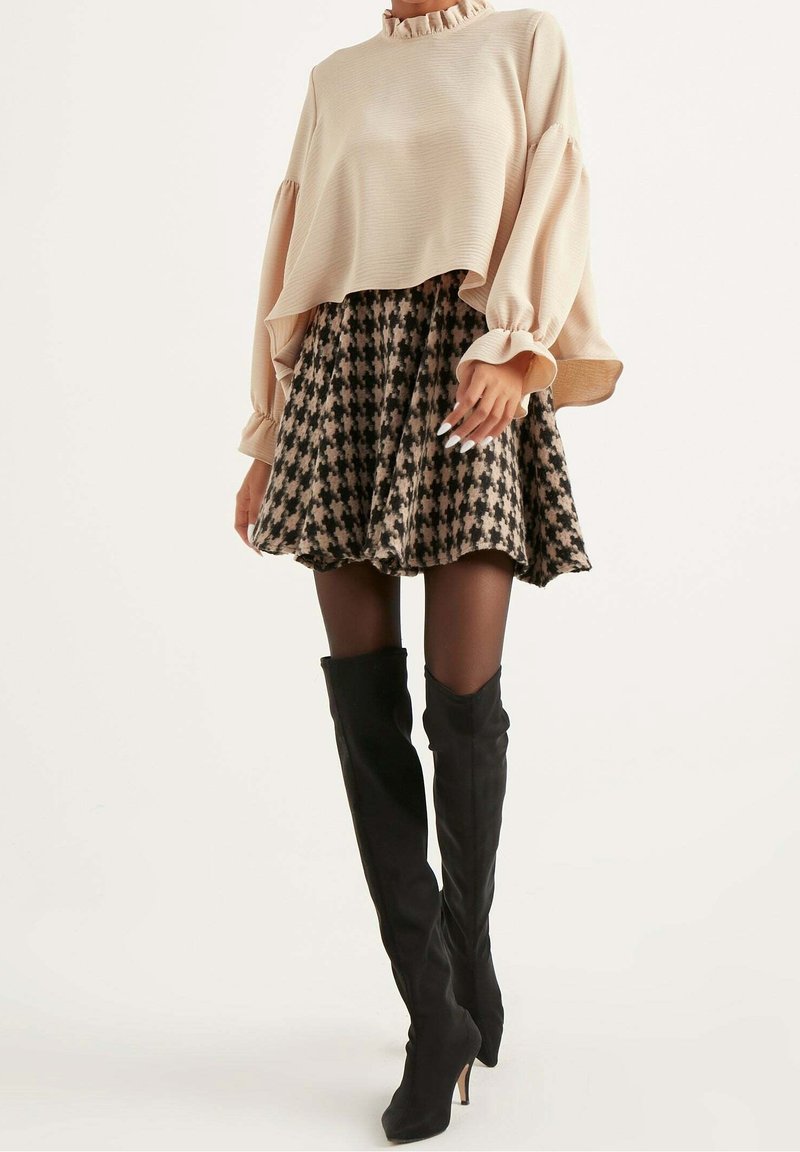 Femme portant un chemisier beige à volants, une jupe pied-de-poule noir et beige, des collants noirs et des bottes à talons hautes noires jusqu'aux genoux.