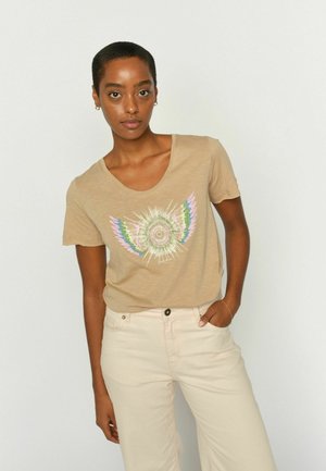 Fiatal nő rövid hajjal, bézs színű mintás pólót és világos bézs nadrágot visel, egyik kezét zsebre téve áll egyszínű háttér előtt.