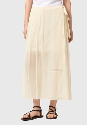 Kremowa długa spódnica z przezroczystą nakładką, ozdobiona wiązaniem z boku, noszona z czarnymi płaskimi sandałami na ramiączkach, na jasnym tle.