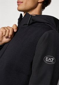 Veste de sport noire avec capuche zippée, tissu texturé et logo EA7 Emporio Armani sur la manche gauche. Design lisse et structuré.
