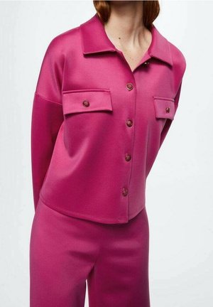 Femme portant une veste boutonnée rose vif avec des poches à rabat, associée à un pantalon assorti, sur un fond blanc uni.