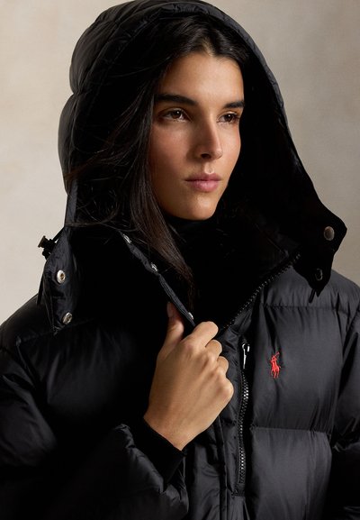 Mujer con chaqueta acolchada negra con capucha, ajustando la cremallera, con el logo rojo de Polo Ralph Lauren visible en el pecho.
