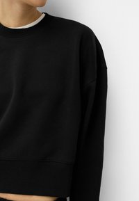 Črna skrajšana športna jopica s krožnim izrezom, rebrastimi manšetami in pasom. Narejena iz gladkega, mehkega materiala z sproščenim krojem.