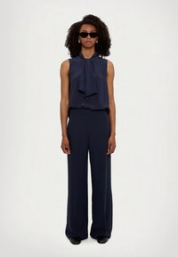 Temno modra brezrokavnik z metuljčkom, kombiniran z visokim pasom in širokimi nogavicami v temno modri barvi. Mat tkanina, minimalističen dizajn in temna sončna očala.