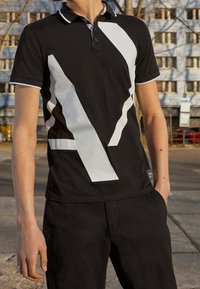 Svart pikétröja med djärv vit grafisk design, kontrasterande krage och ärmsnoddar. Tillverkad av slät tyg, sittande tätt runt torson.