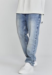Jeans de mezclilla en un azul claro con un corte relajado, que presentan parches descoloridos y un detalle de costura oscura. Combinados con zapatillas blancas.