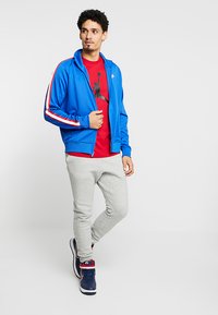 Blå zip-jakke med rød og hvid striber, rød T-shirt med grafik, grå joggers og marineblå sneakers med røde detaljer.