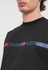 Μαύρο φούτερ με στρογγυλή λαιμόκοψη, με το λογότυπο "TOMMY HILFIGER" σε μπλε και κόκκινες οριζόντιες ρίγες στο στήθος.