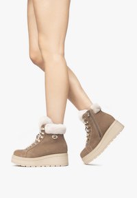 Tan-farbene Wildleder-Stiefeletten mit Fellbesatz, Schnürdesign, goldenen Akzenten und einer dicken, crèmefarbenen Plateausohle, die an den Beinen eines Models gezeigt werden.