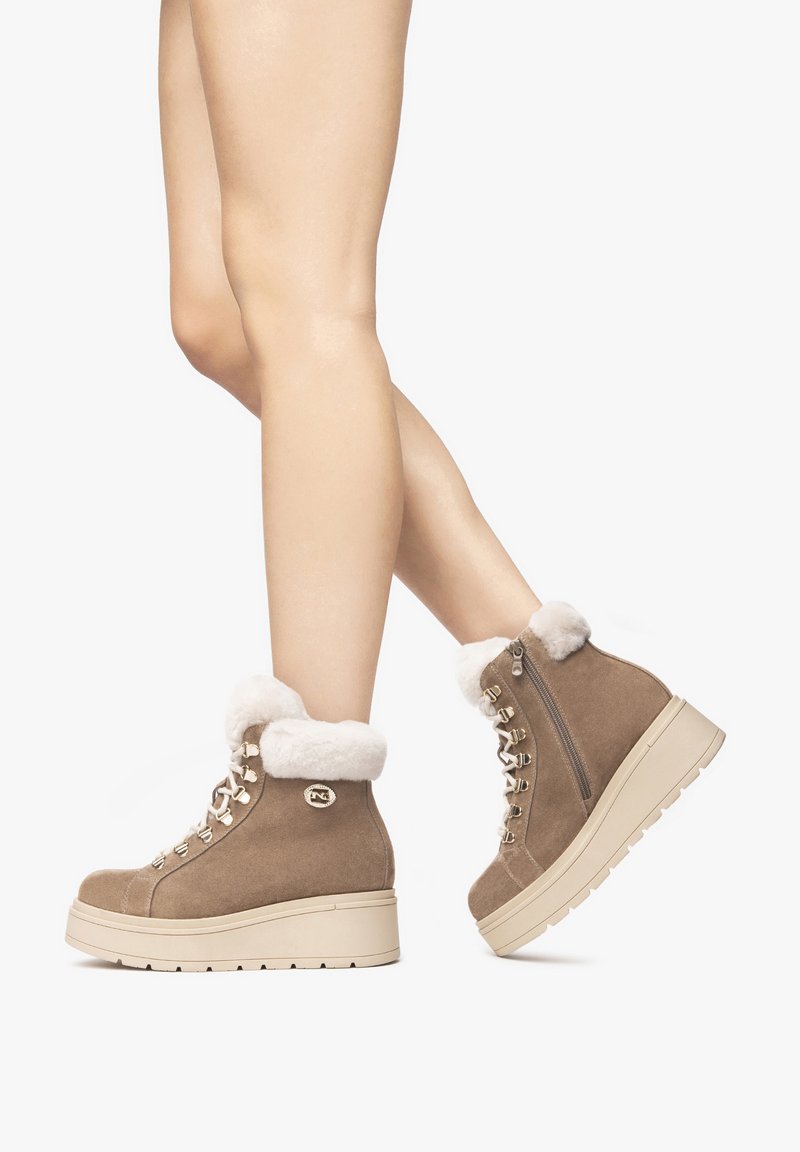 Tan-farbene Wildleder-Stiefeletten mit Fellbesatz, Schnürdesign, goldenen Akzenten und einer dicken, crèmefarbenen Plateausohle, die an den Beinen eines Models gezeigt werden.