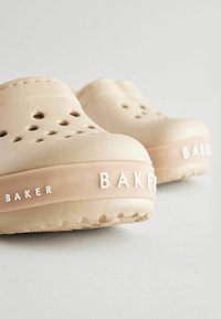 Zuecos beiges con agujeros de ventilación, un acabado texturizado y una suela de goma. "BAKER" está grabado en blanco en los laterales.