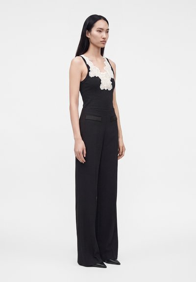 Femme aux longs cheveux noirs debout, portant un haut noir sans manches avec un détail en dentelle blanche et un pantalon noir large taille haute.