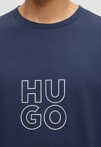 Camiseta de algodón azul marino con letras blancas grandes que dicen "HUGO" en una fuente moderna y cuadrada. Cuello redondo y mangas cortas.