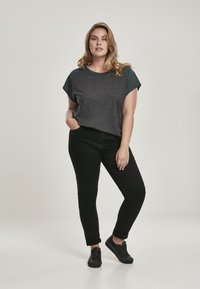 Donkergroen kort mouwen top met groene accenten, gecombineerd met zwarte slim-fit jeans en zwarte sneakers. Model staat met één hand in de zak.