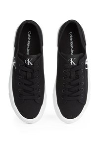 Črni nizki platneni supergi z belimi gumijastimi podplati, črnimi vezalkami in oznakami z logotipom "CK" na jezičku in stranskih panelih.