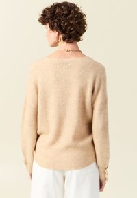 Pull beige à côtes avec une coupe décontractée et un large col. Il se caractérise par un tricot texturé et des poignets retroussés, porté sur un pantalon blanc.