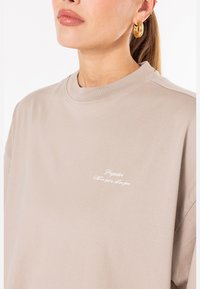 Beige bomulls T-skjorte med rund hals, med små hvite broderte bokstaver på brystet. Glatt tekstur og avslappet passform.
