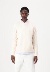 Pull crème clair avec un col rond, en tissu texturé, avec des poignets côtelés, superposé sur une chemise rayée bleue et blanche, associé à un pantalon blanc.