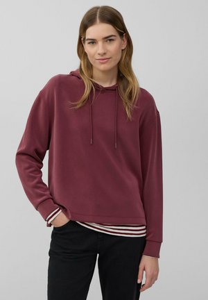 Jeune femme avec des cheveux châtain clair portant un hoodie bordeaux et une chemise rayée, se tenant avec une main dans la poche contre un fond uni.