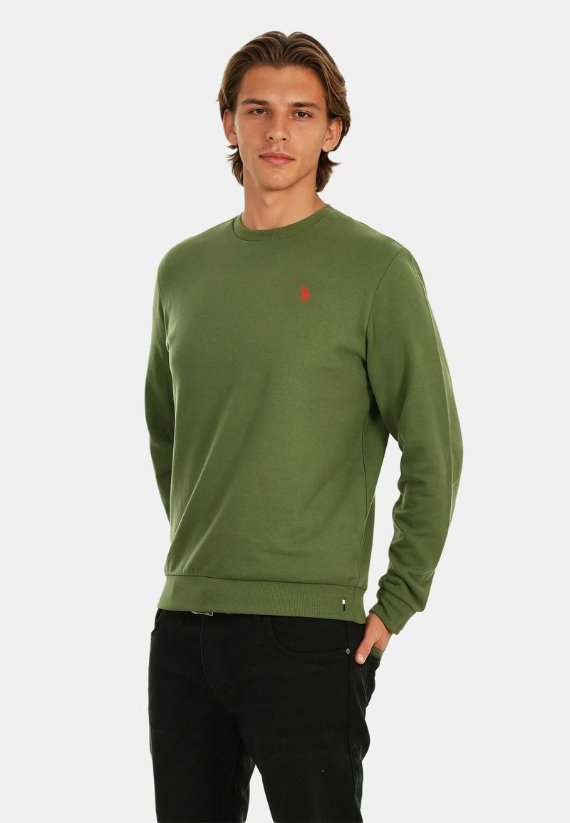 Green cotton sweatshirt with ribbed cuffs and hem, featuring a small red logo on the chest. Model wears black pants. Simple design.