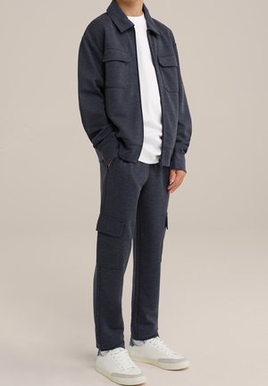 Granatowa kurtka o fakturze z dwiema kieszeniami na piersiach oraz dopasowane spodnie cargo. Biała koszulka pod spodem i białe sneakersy dopełniają cały strój.