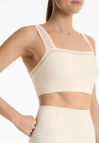 Cream-colored sports bra with square neckline, wide straps featuring cutout pattern, smooth texture, and seamless finish.