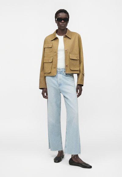 Γυναίκα με μαύρα γυαλιά ηλίου, μαύρη κοντή μπεζ ζακέτα με μεγάλες τσέπες, λευκό τοπ, ανοιχτό μπλε τζιν παντελόνια με ψηλή μέση και μαύρα φλατ παπούτσια.