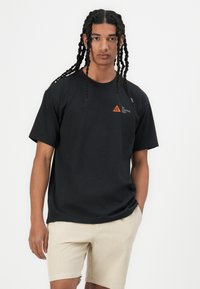 Zwarte katoenen t-shirt met korte mouwen en een klein oranje driehoeklogo. Gecombineerd met beige shorts die een ontspannen pasvorm en textuur tonen.