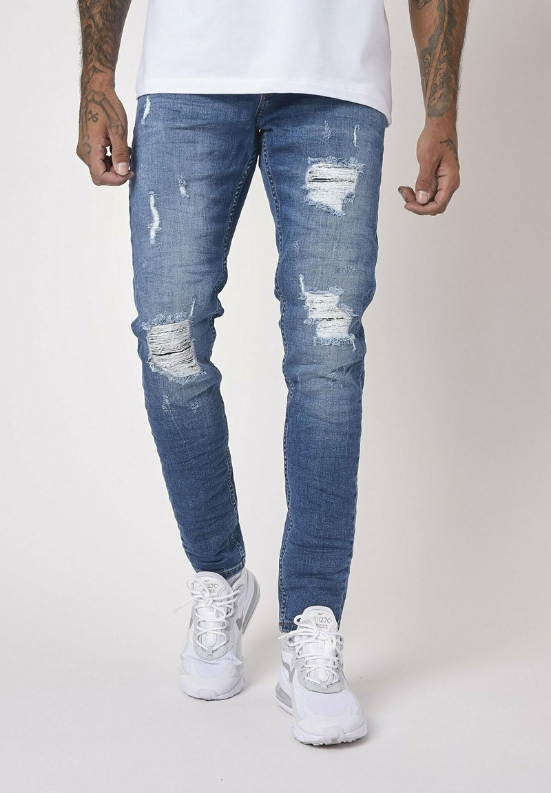 Blue denim jeans with ripped detailing and a slim fit, paired with white sneakers. Features fading and frayed edges for a distressed look.