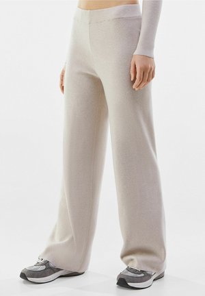 Personne portant un pantalon en maille beige à jambes larges et des baskets grises, debout devant un fond clair uni.